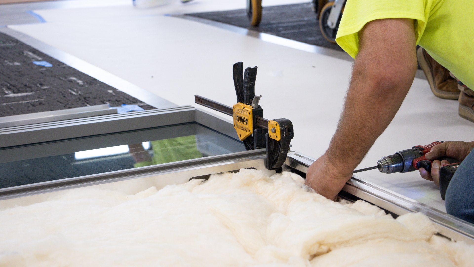 A worker installs insulation into an architectural wall panel using a drill and clamp.