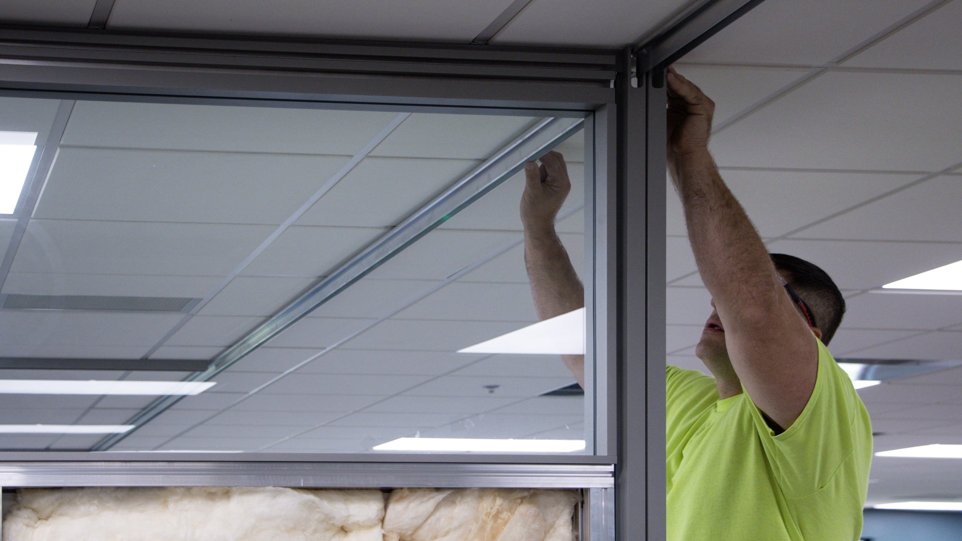 A worker secures the top frame of an architectural wall panel inside a finished office ceiling.